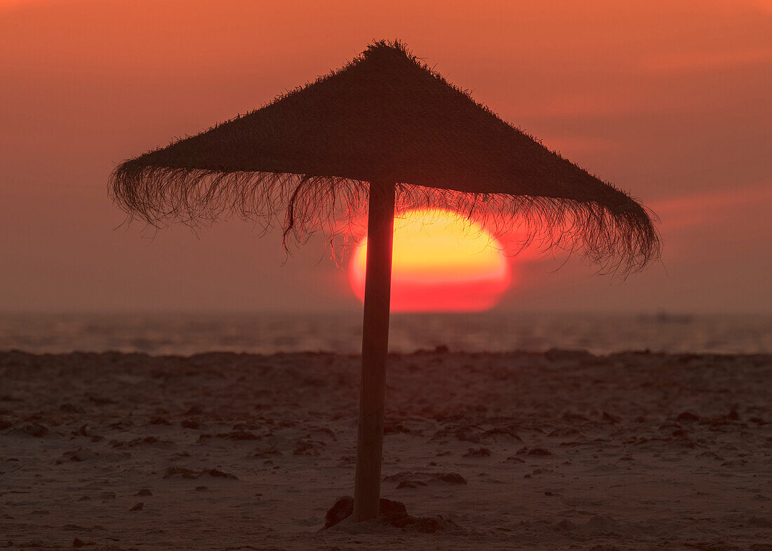 Silhouette of a beach umbrella on the beach with a colourful sun sinking into the horizon over the ocean, Tarifa, Cadiz, Andalusia, Spain