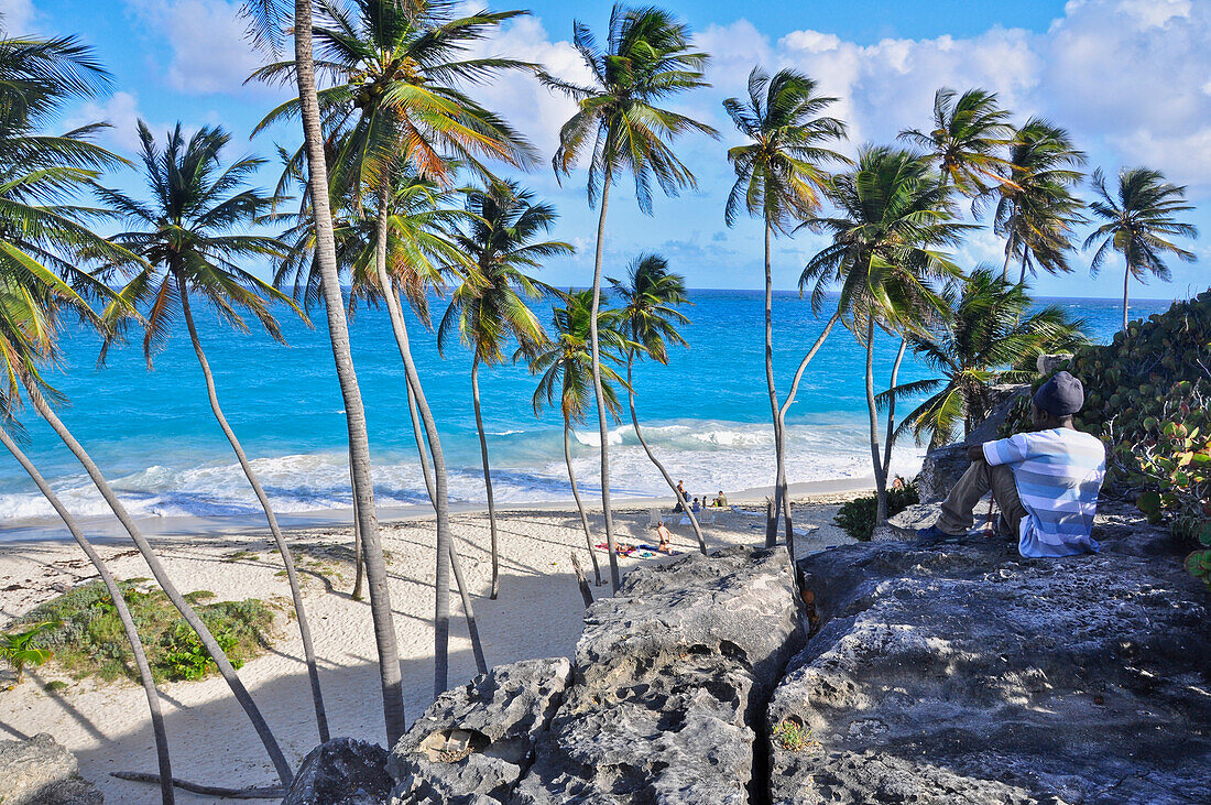 Einheimischer junger Mann auf Felsen an tropischem Strand mit Palmen, Meer, Bottom Bay, Südküste, Barbados, Kleine Antillen, Westindische Inseln, Windward Islands, Antillen, Karibik, Mittelamerika