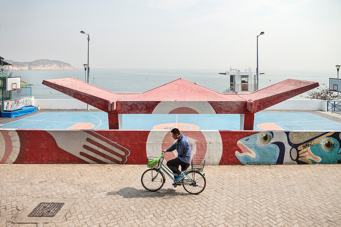 Chinese auf Fahrrad fährt an Sportplatz vor Meer vorbei, Insel Cheng Chau, Hongkong, China, Asien