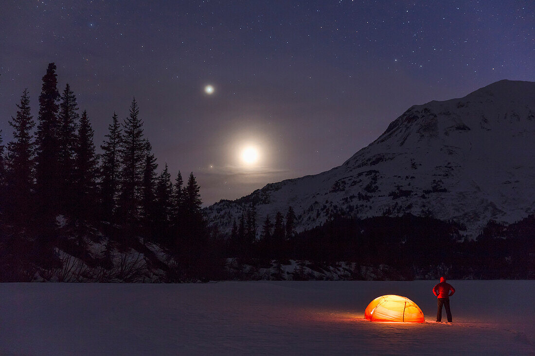 Nighttime view of a man standing next to a lit tent on a snow covered Trail Lake with the moon, stars, and planets overhead, Moose Pass, Kenai Peninsula, Southcentral Alaska