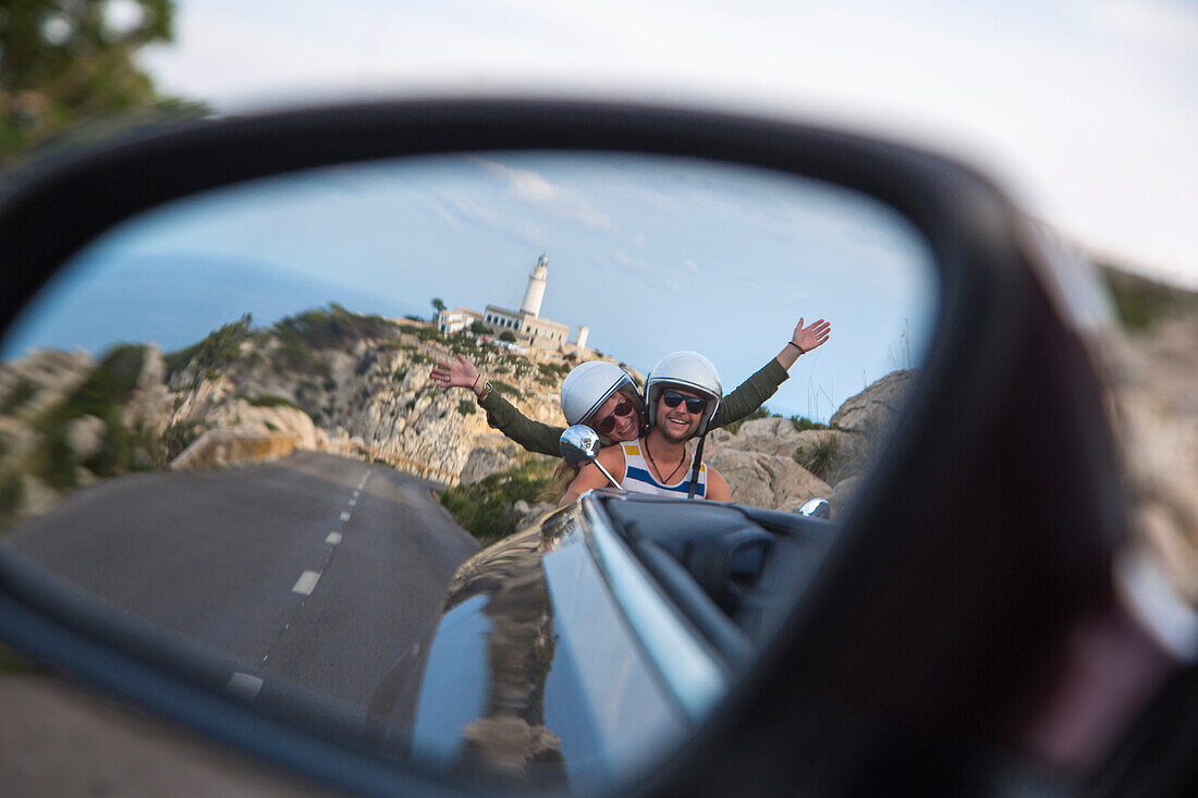 Blick in den Rückspiegel eines Sunny Cars Cabriolets auf junges Paar auf rotem Vespa Motorroller auf Straße entlang der Halbinsel Cap de Formentor mit Leuchtturm Faro de Formentor im Hintergrund, Cap de Formentor, Palma, Mallorca, Balearen, Spanien