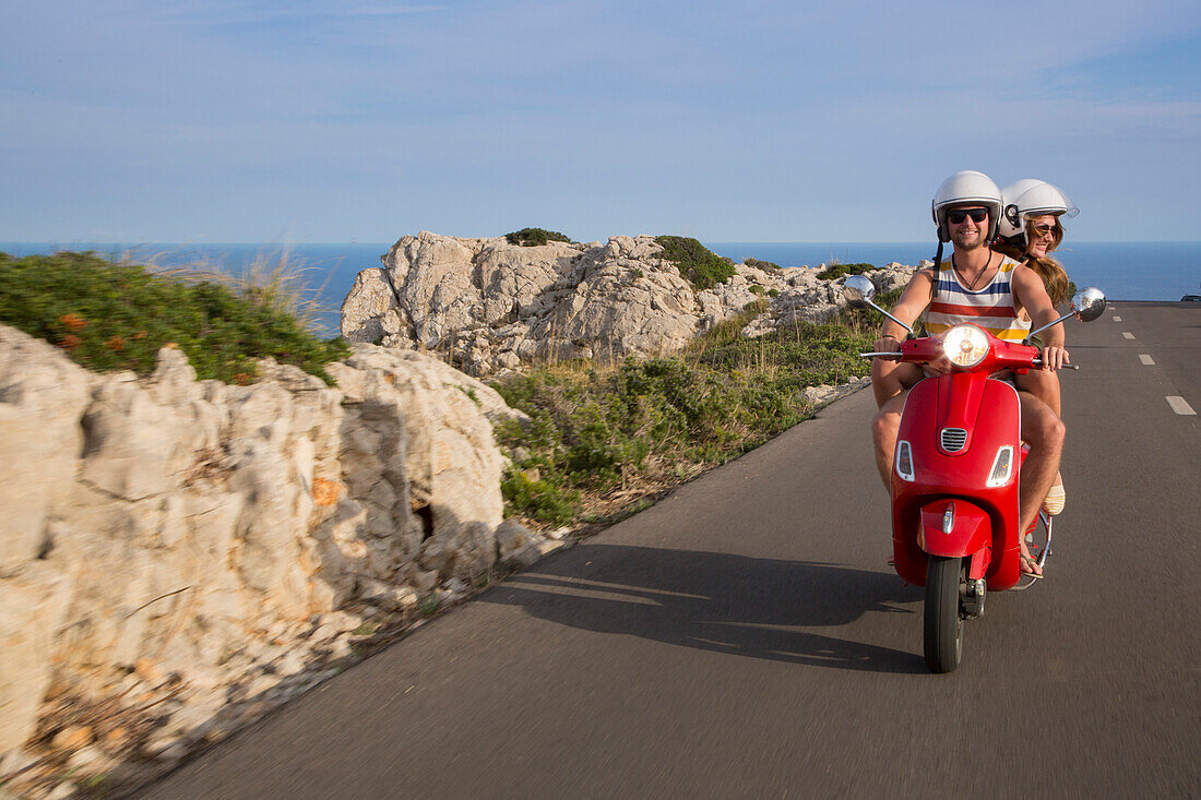 Junges Paar fährt roten Vespa Motorroller auf Straße entlang der Halbinsel Cap de Formentor mit Meer im Hintergrund, Cap de Formentor, Palma, Mallorca, Balearen, Spanien