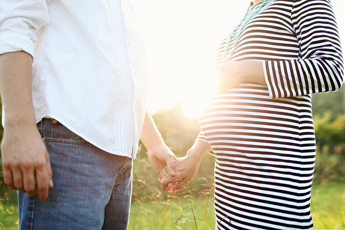 Pregnant Caucasian couple holding hands in field