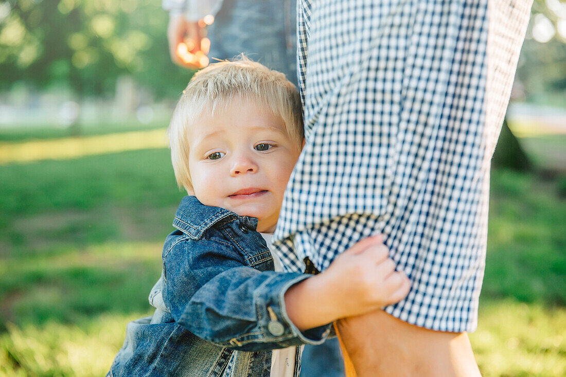 Boy clutching leg of father in grass in park