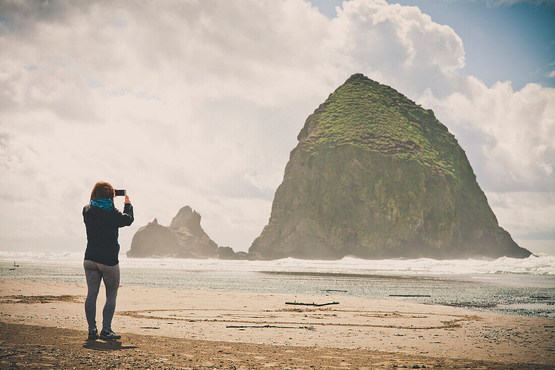 A young woman uses her smartphone to take a picture of Haystack Rock on a cloudy day while visiting Cannon Beach, Oregon.