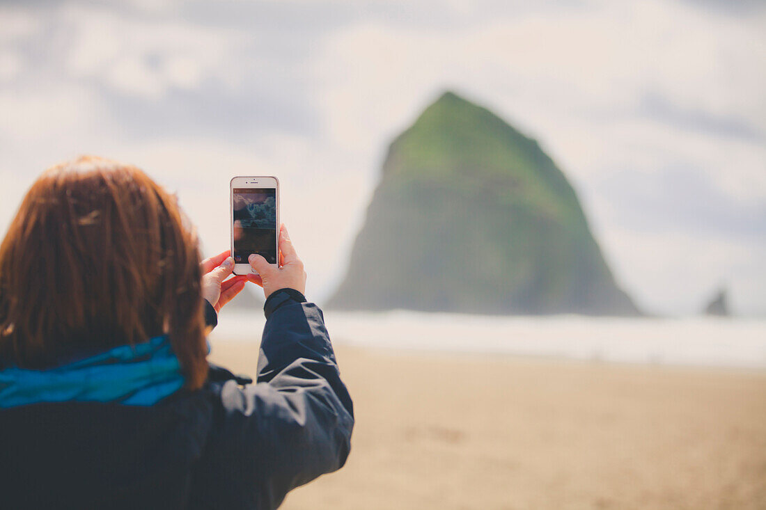 A young woman uses her smartphone to take a picture of Haystack Rock while visiting Cannon Beach, Oregon.