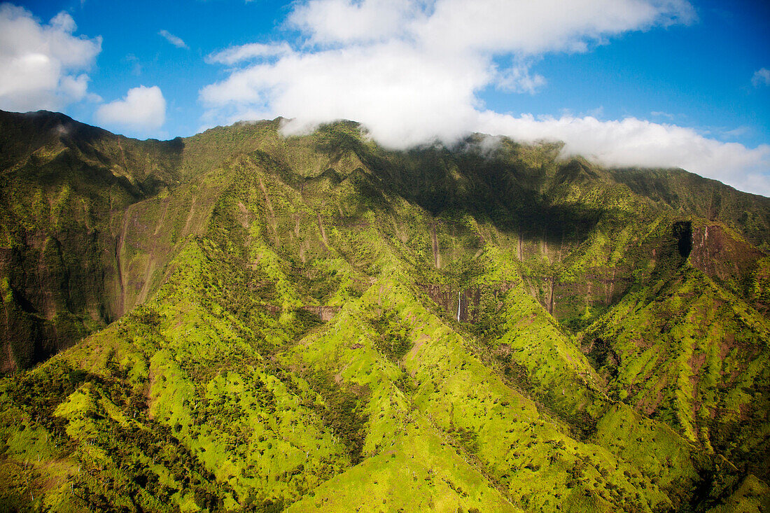 Aerial image of lush green mountain slopes under soft clouds and blue sky.