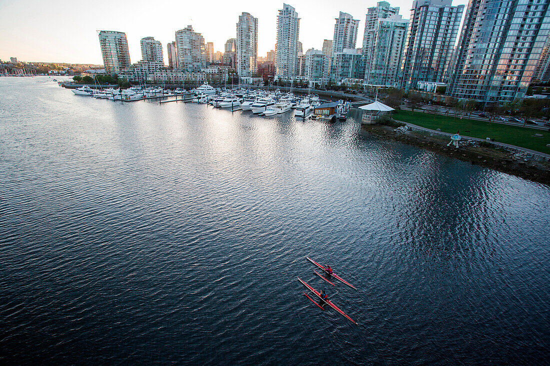 Two rowers paddle through calm water with Vancouver skyline in distance, shot from a bridge.