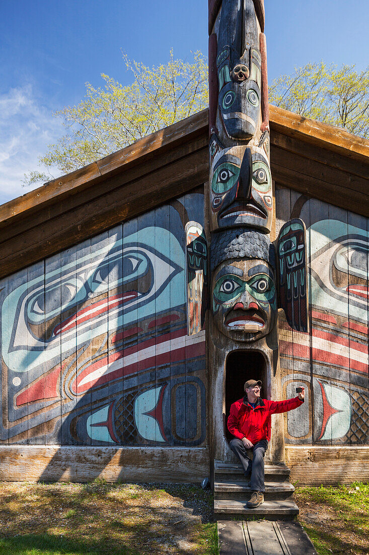Man sits on steps of a Tlingit clan house with smart phone, Totem Bight Historic State Park, Ketchikan, Southeast Alaska, USA, Spring