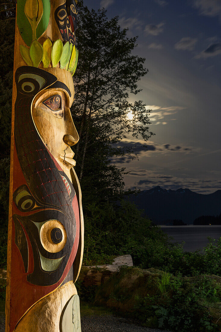 A large totem pole lit up at night in Sitka National Historic Park with the moon and clouds in the background, Sitka, Alaska, United States of America