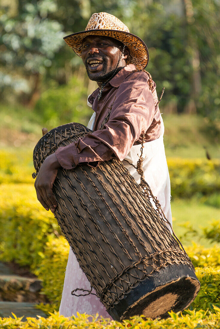 An African man in a brown shirt and pink and white patterned wrap with a leopard-print hat plays a drum, Nkuli, Western Province, Rwanda