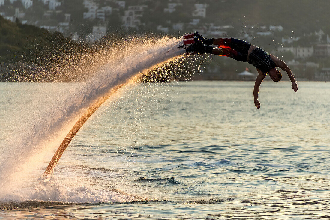 Silhouette of Flyboarder diving in perfect high backlit arc in the late afternoon sunshine with a village in the background, Torba, Mugla Province, Turkey