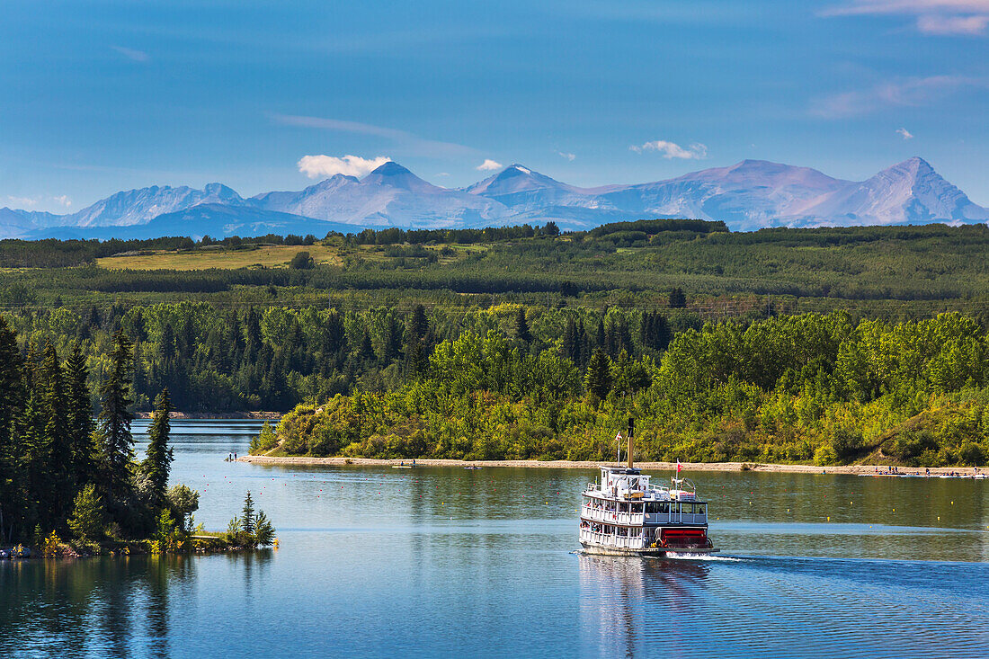 Paddlewheel boat on lake with tree lined shoreline, hills and mountains in the background and blue sky and clouds, Calgary, Alberta, Canada