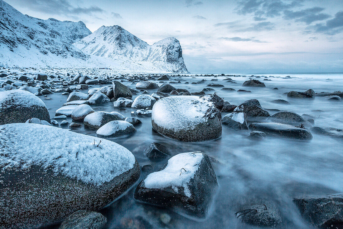 Rocks in the cold sea and snow capped mountains under the blue light of dusk, Unstad, Lofoten Islands, Arctic, Northern Norway, Scandinavia, Europe