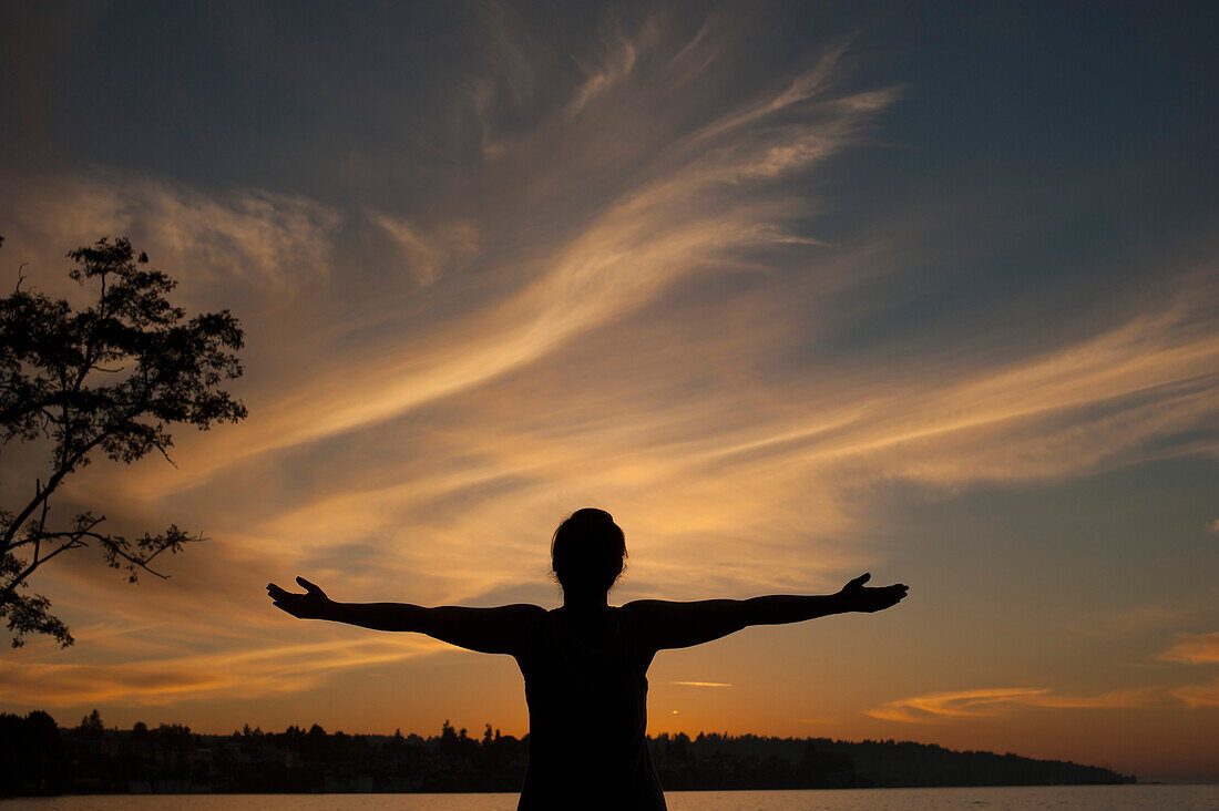 Silhouette young woman standing with arms outstretched on lakeshore during sunset