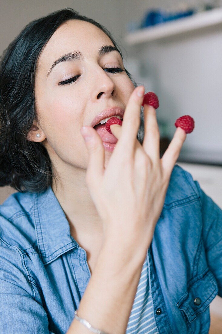 Young woman eating raspberries on fingers at home