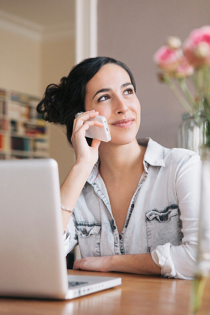 Beautiful young woman using mobile phone with laptop on table at home