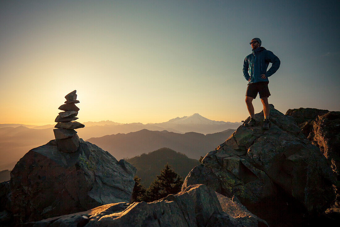 A hiker stands on a large rock, opposite the summit cairn or Sauk Mountain in the North Cascade Mountain Range, Washington.