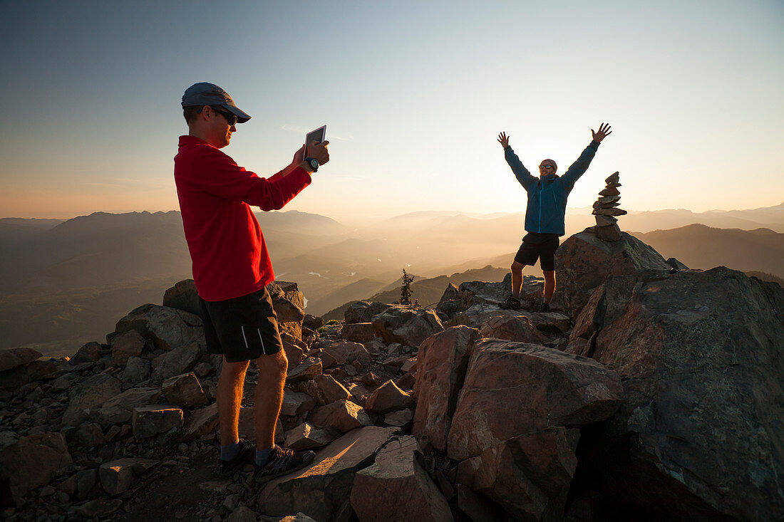 A hiker uses a tablet to take a picture of his friend standing next to the summit cairn of Sauk Mountain, Washington.