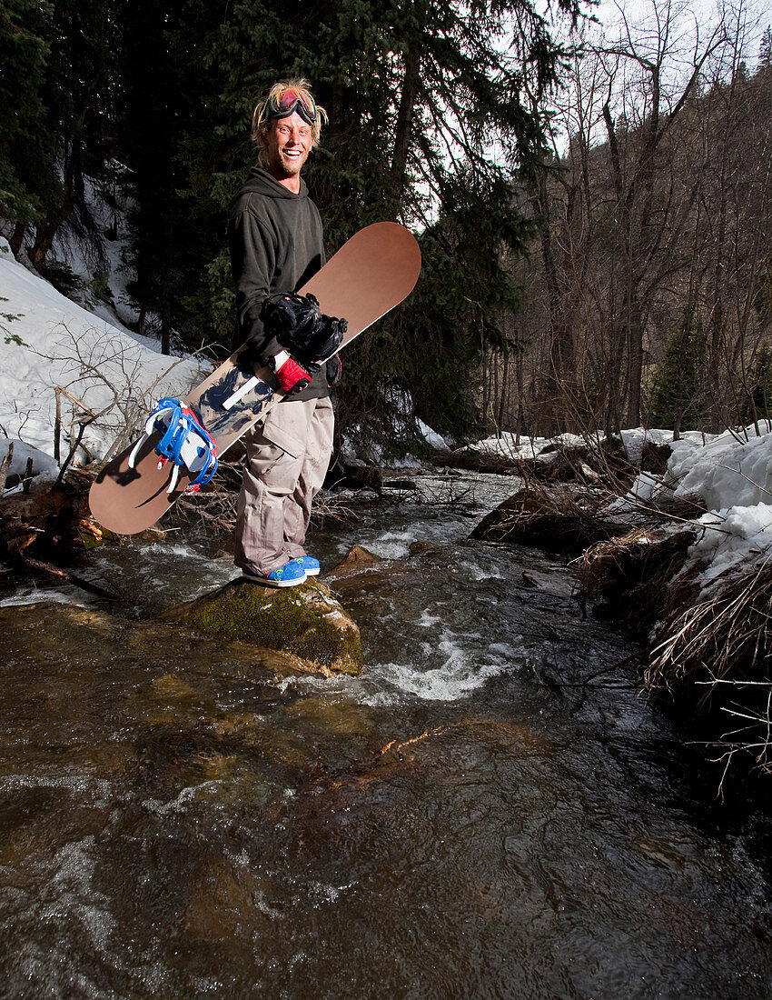 A snowboader with blonde hair stands on a rock in the middle of a creek while holding his board in his hand.
