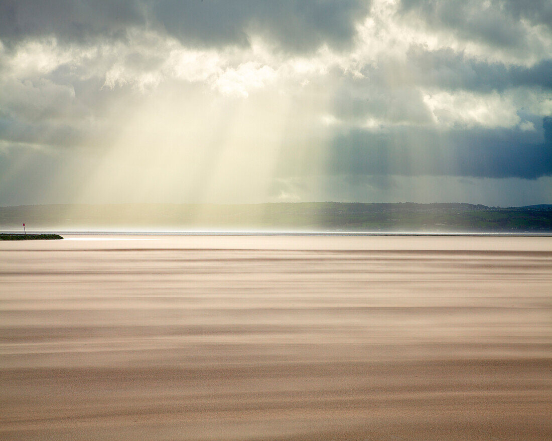 Crepuscular rays through a stormy sky while shifting sands create a cloud underfoot as wind whistles across the beach, West Kirkby, Wirral, England, United Kingdom, Europe