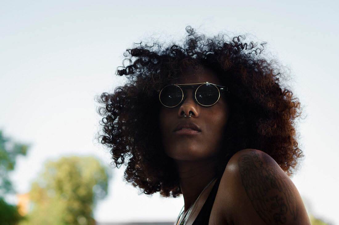 Portrait einer jungen afroamerikanischen Frau mit Sonnenbrille in der Natur und blauem Himmel, München, Bayern, Deutschland