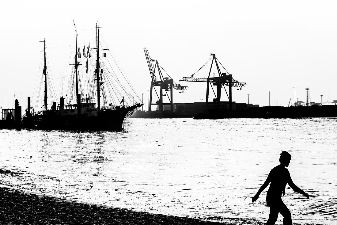 Silhouette einer Frau die in Oevelgönne vor der Kulisse des Museumshafens und dem Containerterminal Burchardkai am Strand spazieren geht, Hamburg, Deutschland