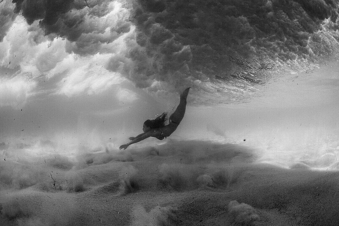 Black and white photo of a girl in a bikini swimming underwater in the ocean.  She goes underneath the waves over a sandy bottom covered by clear water.