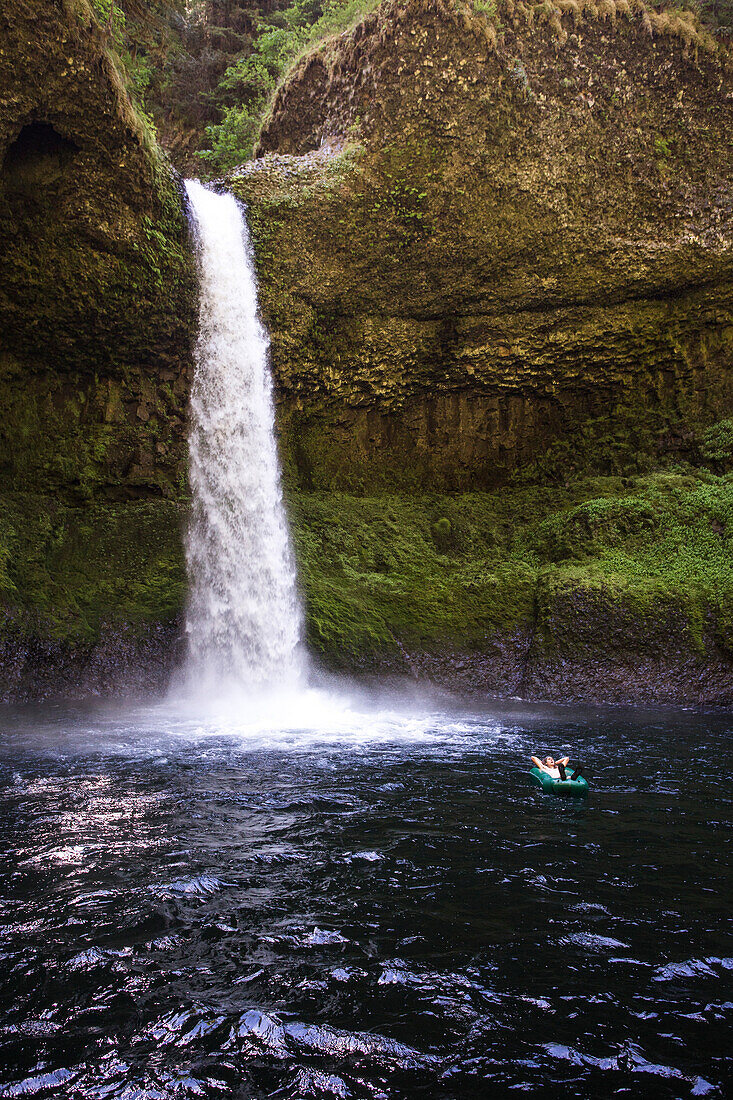 A man floats in a one-person pack raft in a dark pool below a tall waterfall.