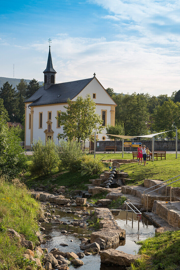 Wasserspiele des Bischofsheimer Was(s)ErlebnisWeg an einem Bach mit katholischer Friedhofskapelle St. Josef dahinter, Bischofsheim, Rhön, Bayern, Deutschland