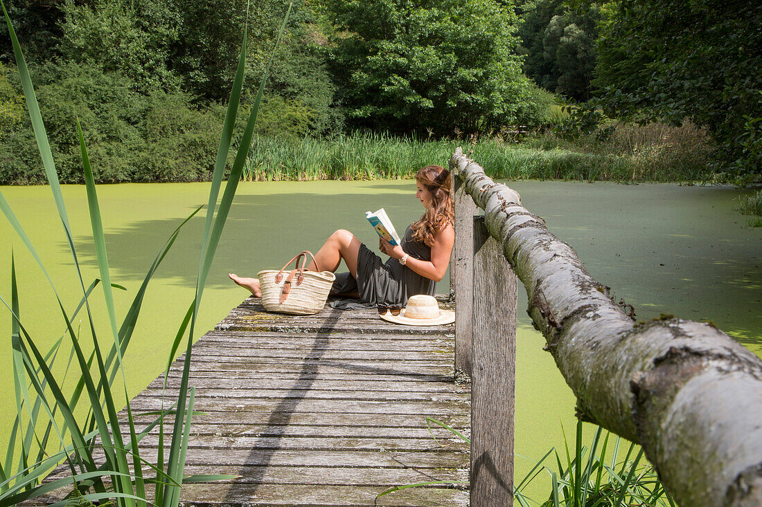 Junge Frau sitzt auf Steg am Teich und liest ein Buch, Haunetal, Rhön, Hessen, Deutschland [MR]