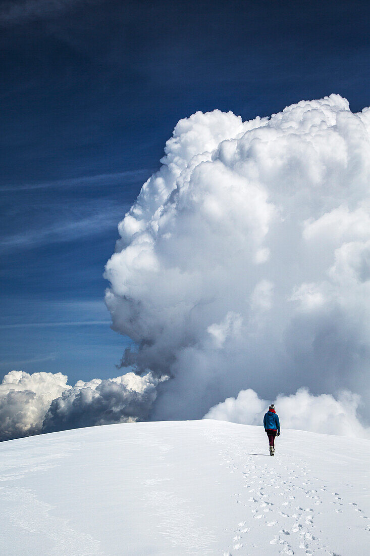 A woman in parka, tights, and ski boots walks across a snowy summit plateau with a large cloud in background.