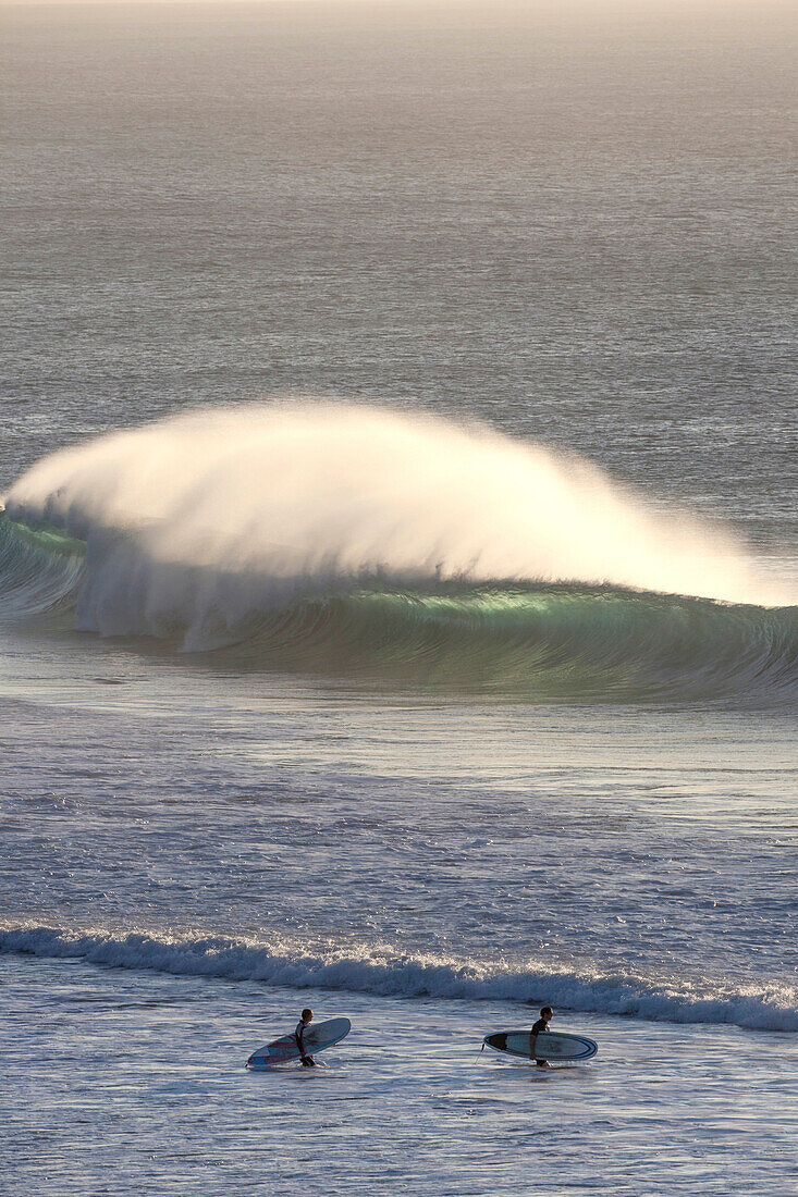 Two surfers on the beach with big waves breaking in the background