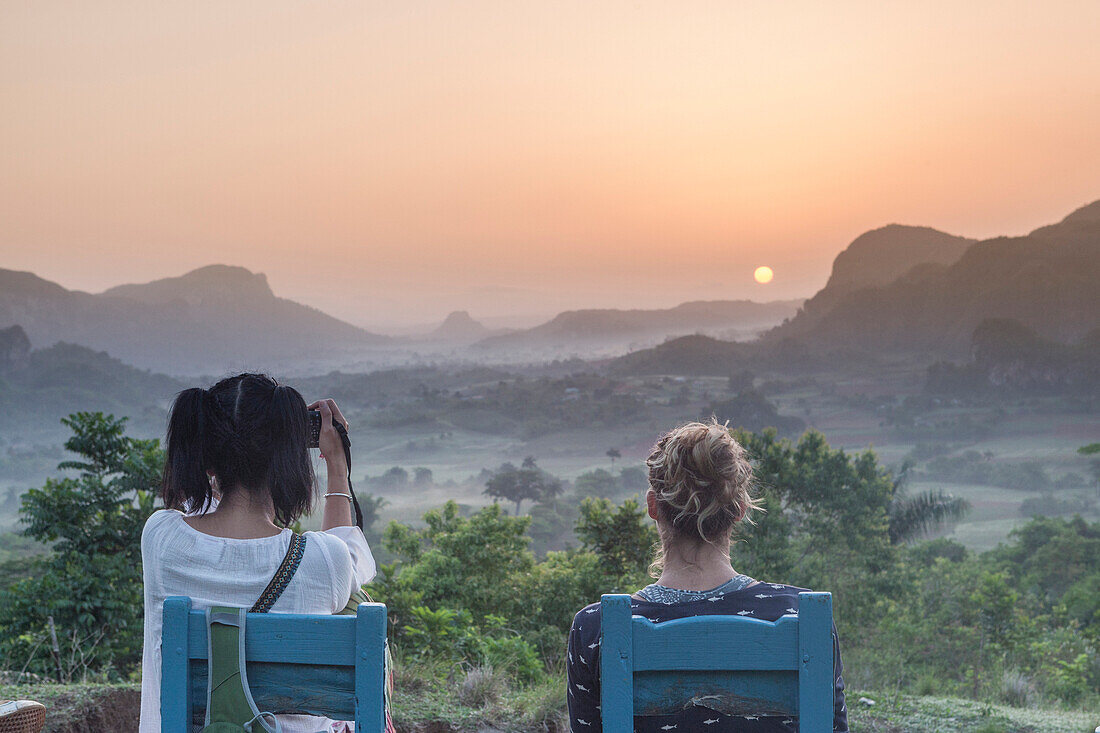 Two girls sitting on wooden chairs watching the sunrise above Vinales National Park from Los Acuaticos, the final point of an early morning trekking famous to watch the sun rising. Vinales, Cuba