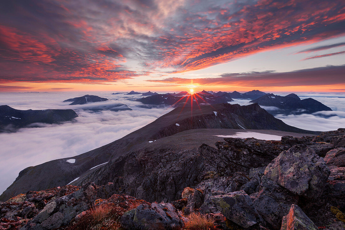 Wide view from top of Skårasalen (1542m) with an impressive sunset over the clouds and the mountains of the Sunnmøre Alps with rocks in foreground, Møre og Romsdal, Norway, Scandinavia