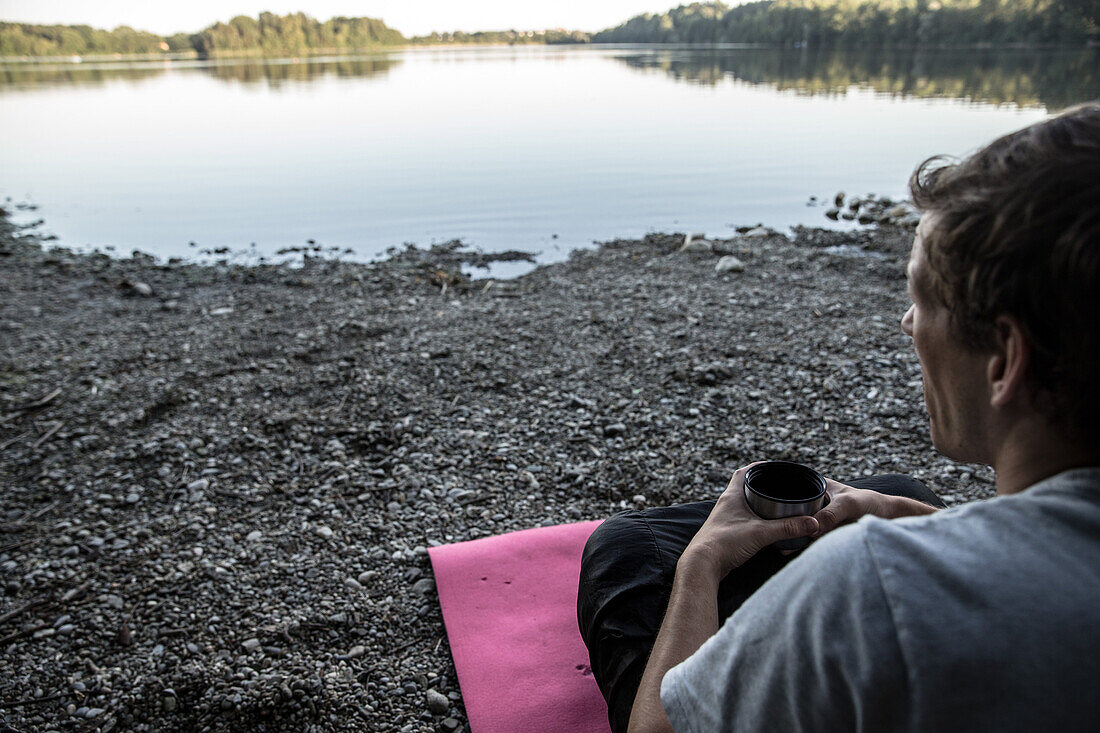 Young man sitting at a lake and drinking coffee, Freilassing, Bavaria, Germany