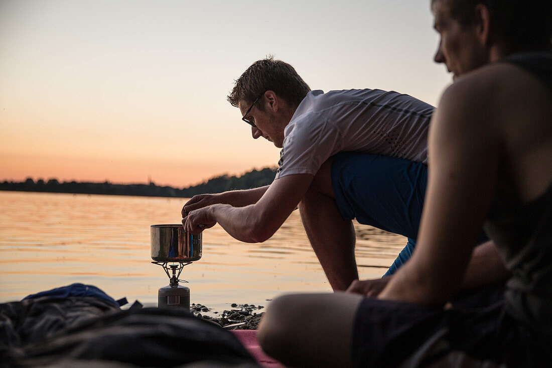 Junger Mann kocht mit einem Campingkocher an einem See, Freilassing, Bayern, Deutschland