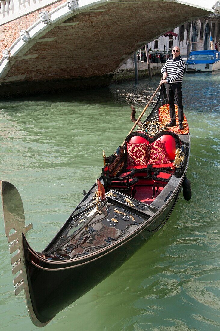 Richly decorated red gondola, with gondolier, on canal, bridge Cannaregio or delle Guglie in the background, Venice, Italy.