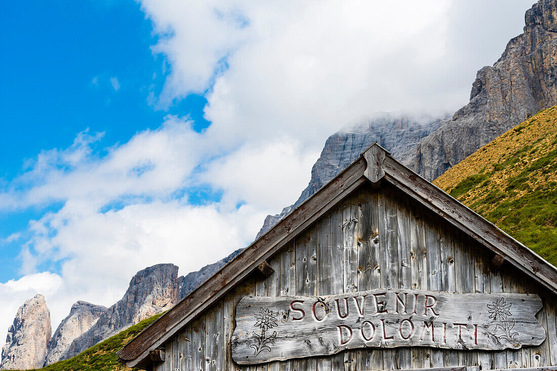 ' A timber house with carved label ''Souvenir Dolomiti'' as a souvenir shop in fornt of the mountain scenery at the Sellajoch, Canazei, the Dolomites, South Tirol, Alto Adige, Italy'