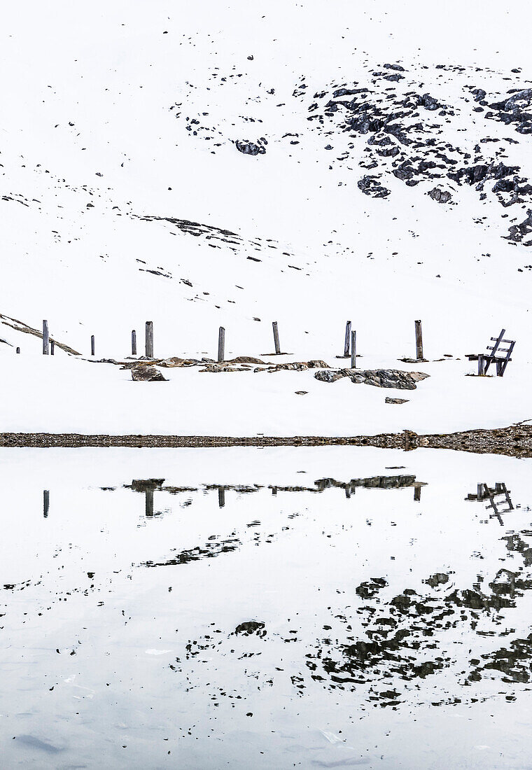A wooden bench and wooden stakes in a snowy scenery are reflected in a lake, Stilfser Joch, Passo dello Stelvio, Trafoi, South Tirol, Alto Adige, Italy