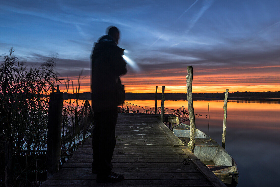Mann steht mit Taschenlampe auf einem Steg des Rangsdorfer Sees bei untergehender Sonne - Deutschland, Brandenburg, Rangsdorf