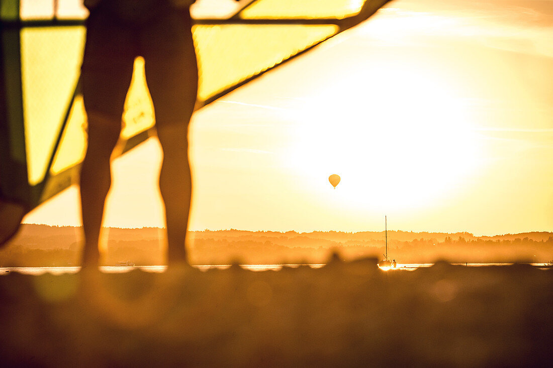 Junger Windsurfer checkt sein Segel und blickt zu einem Heißluftballon an einem sonnigen Abend, Ammersee, Bayern, Deutschland
