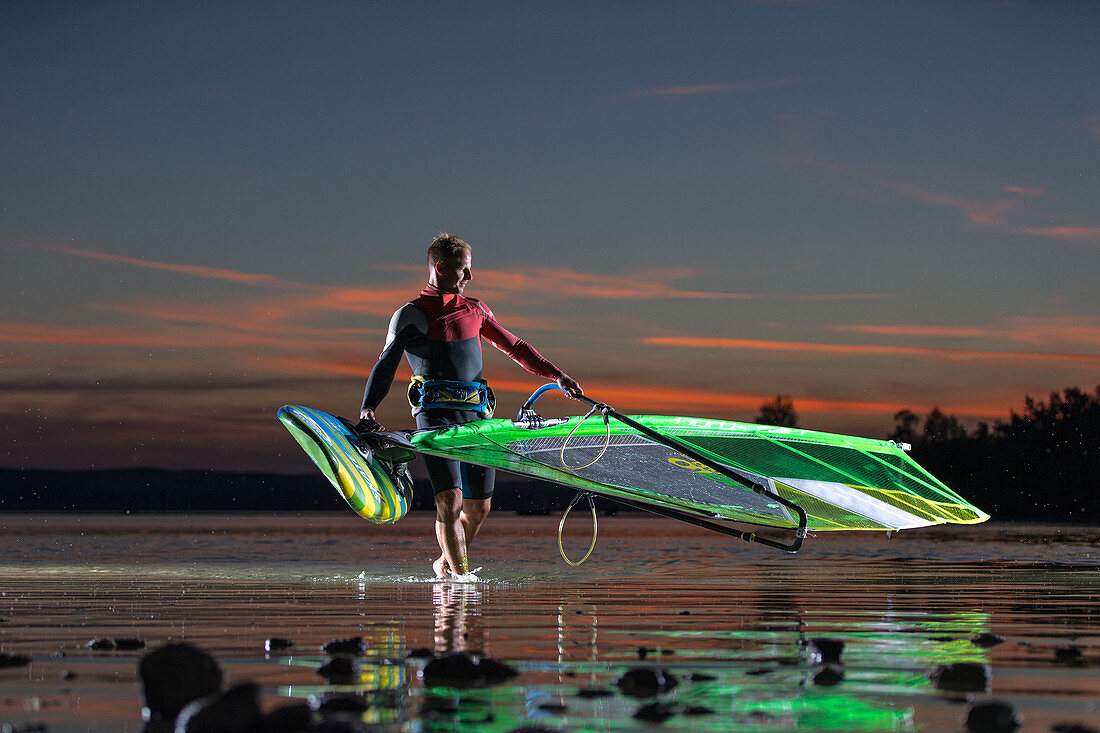 Young male windsurfer bringing his board and sail out of the water while sunset, Ammersee, Bavaria, Germany