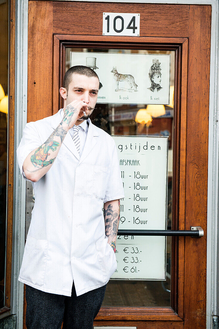 Rotterdam, Netherlands. Barber store Schorem. Portrait of a barber with tattoos on his arm, smoking a cigarette just outside the front door.