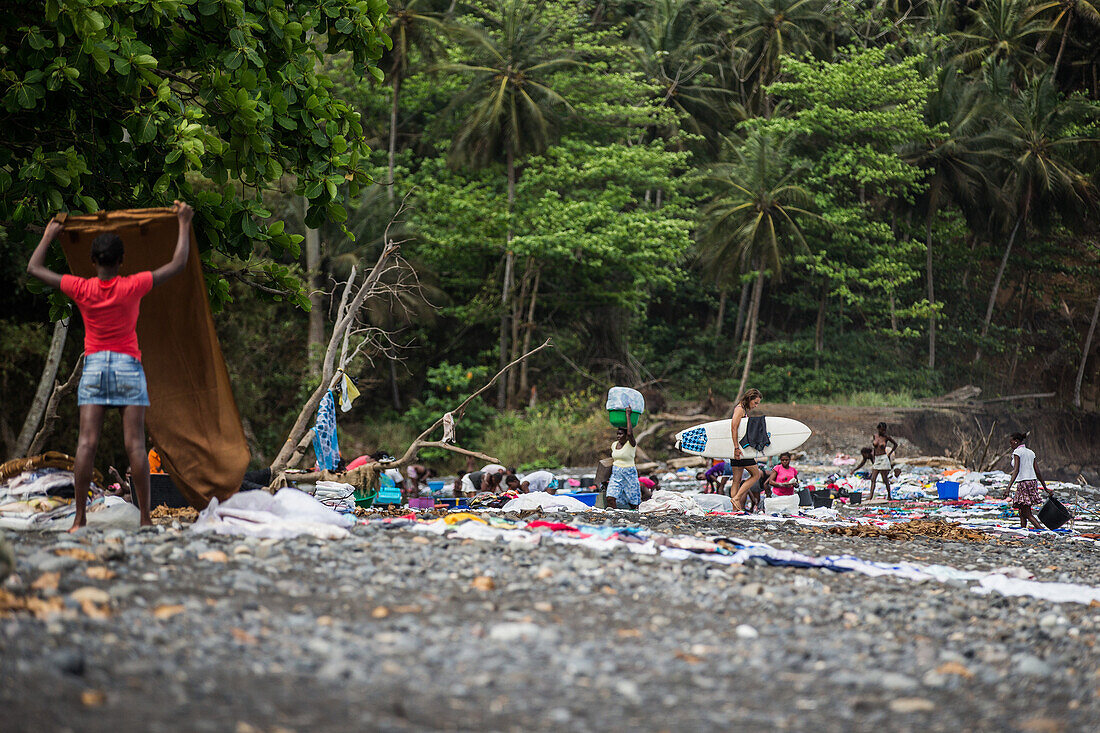 Young female surfer carrying her board to the sea between many local people, Sao Tome, Sao Tome and Principe, Africa