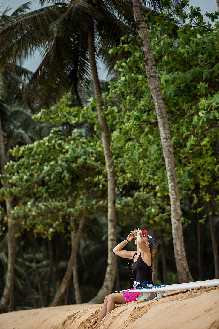 Young female surfer sitting on the beach and drinking from a coconut, Sao Tome, Sao Tome and Principe, Africa