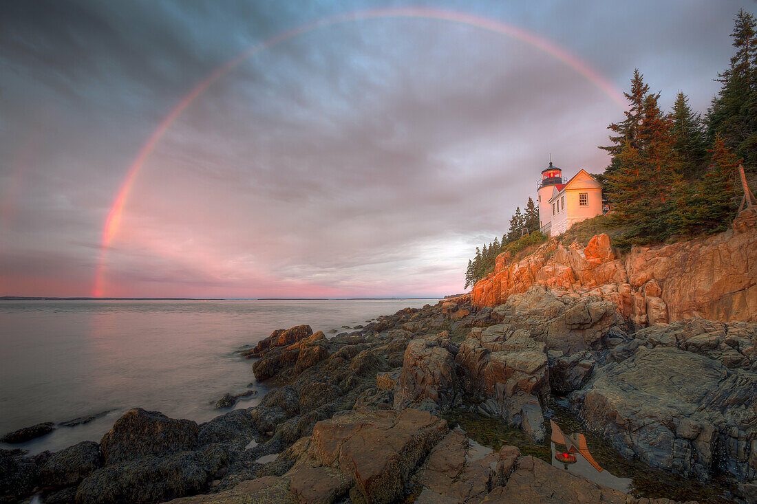 A double rainbow appears briefly at sunrise over Bass Harbor Head Lighthouse, which is reflected in a tidal pool as it overlooks the entrance to Bass Harbor and Blue Hill Bay in Tremont, Maine.