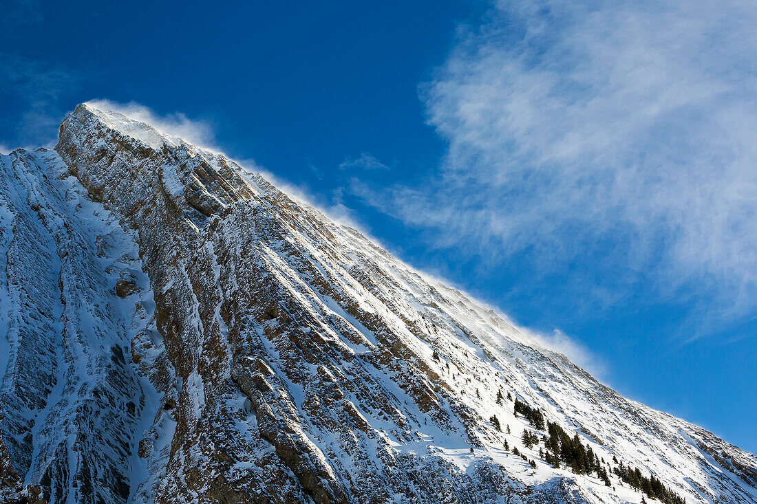 'Close up of a snow covered mountain peak with wind blowing snow off the slope with blue sky and clouds; Kananaskis Country, Alberta, Canada'