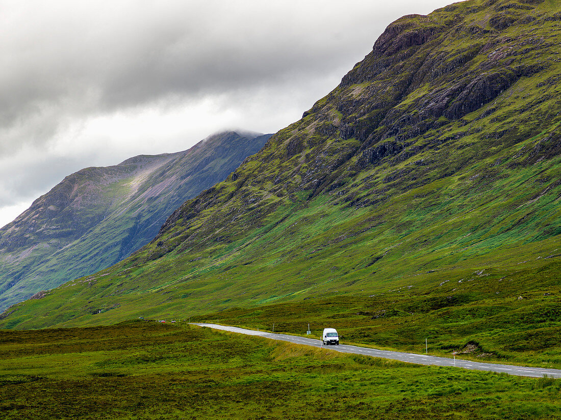 'A vehicle travels on a road beside rugged mountains under a cloudy sky; Scotland'