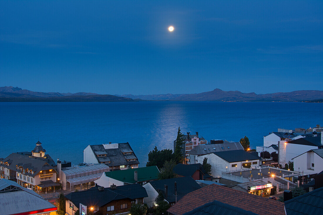 'The moon rising over a lake from above the town roof tops showing some yellow city lights against the blue scene; Bariloche, Argentina'